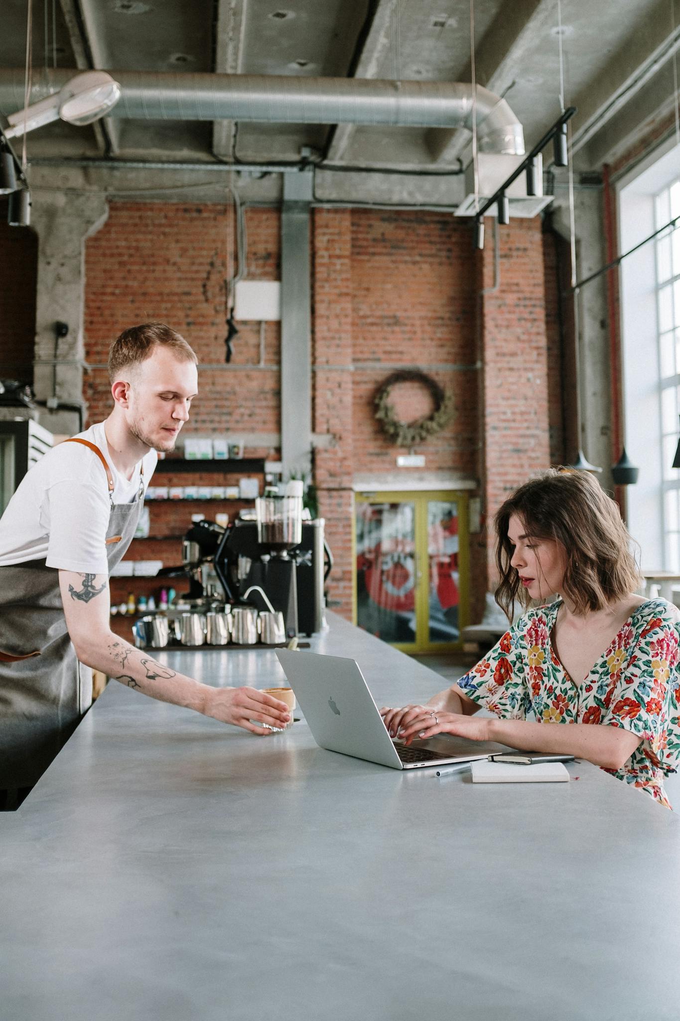 Barista serves a drink to a woman working on a laptop in a modern café.